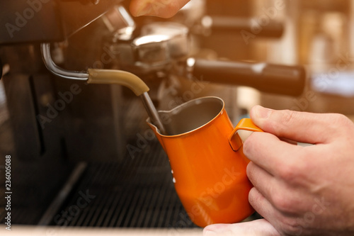 Male barista using coffee machine for steaming milk in pitcher, closeup