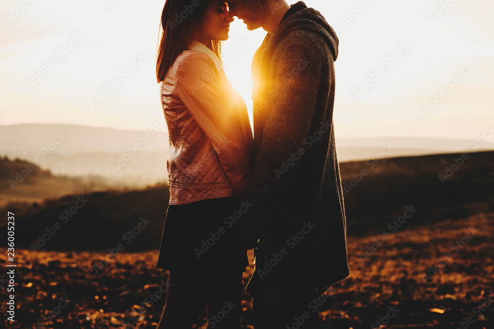 Side view of crop young man and woman touching noses while standing on background of amazing countryside and beautiful sundown sky.Crop couple against sunset sky - obrazy, fototapety, plakaty 