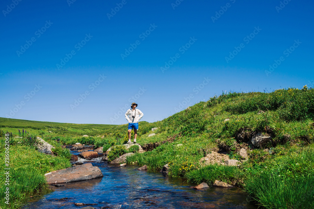 Obraz premium Traveler with camera on stone in mountain creek. Adventure of tourist. Hiking in mountains. Rich vegetation of highlands. Stream of clear water in brook. Vivid sunny landscape of majestic nature.