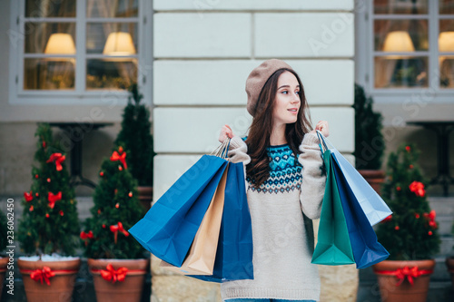 Outdoor portrait of smiling woman posing on street, holding paper shopping bags, looking at camera, wearing stylish clothes. Winter holidays, Christmas, New Year concept.