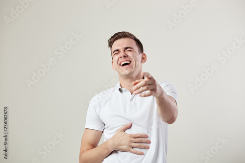 Guy dressed in a white t-shirt and jeans laughs and keeps hand on the belly on a white background in the studio photo