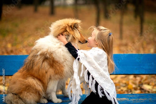 A girl in a white poncho is sitting on a bench in the autumn park and kiss a red collie dog