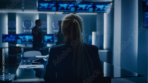 Female Special Agent Works on a Laptop in the Background Special Agent in Charge Talks To Military Man in Monitoring Room. In the Background Busy System Control Center with Monitors Showing Data Flow. photo