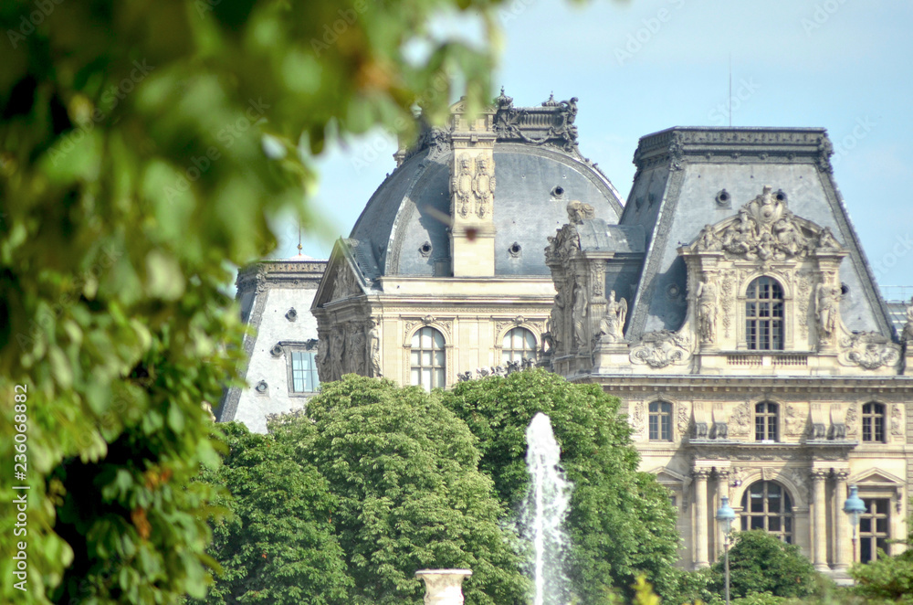 The classical buildings of the Louvre are framed by the trees of the Tuileries Gardens. A fountain is in the foreground. The sky is blue.