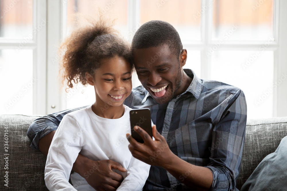 Happy African American family having fun together, funny father with stuck out tongue and little daughter make selfie photo, watching video, sitting on couch at sitting room, using mobile phone