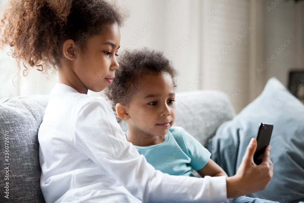 Pretty African American children using smartphone at home, little preschooler girl sitting together on couch with toddler boy holding phone in hands, taking photo, making selfie, watching video Pretty African American children using smartphone at home, little preschooler girl sitting together on couch with toddler boy holding phone in hands, taking photo, making selfie, watching video