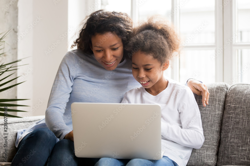 Happy African American attractive mother and little preschooler daughter using laptop, sitting together on couch, sofa in living room at home, watching video, shopping online, looking at screen Happy African American attractive mother and little preschooler daughter using laptop, sitting together on couch, sofa in living room at home, watching video, shopping online, looking at screen