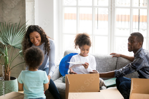 Happy large African American family unpacking cardboard boxes with belongings, just moving, sitting together on couch, little daughter and toddler son helping parents with packaged things, new home