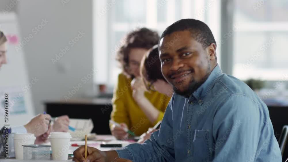 Tilt down shot of joyous African American businessman in denim shirt writing with pencil and then looking at camera and smiling while group of colleagues talking in background Tilt down shot of joyous African American businessman in denim shirt writing with pencil and then looking at camera and smiling while group of colleagues talking in background