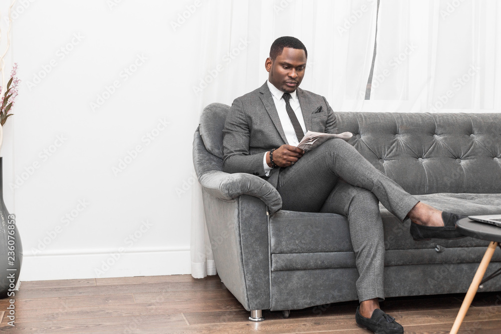 Young African American businessman in a gray suit reading a newspaper while sitting on a sofa.