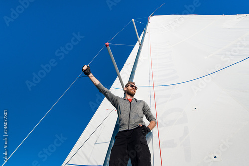 Young man stands on yacht boom and looks forward. He holds on mast rope with hand. Young man poses. He wears grey hoody and black pants.