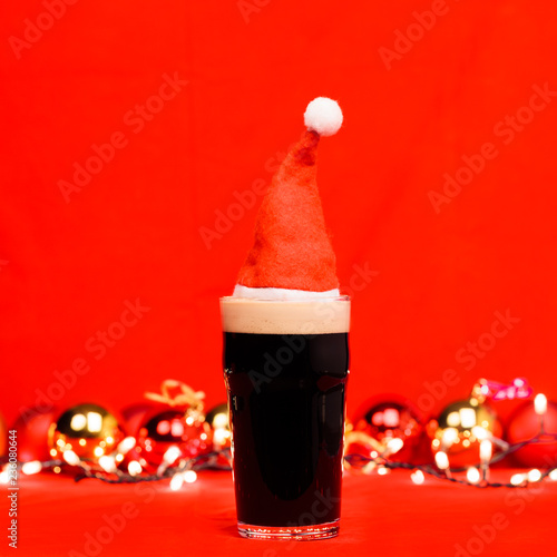 Dark ale beer in pint glass with red santa hat christmas baubles and lights on red background photo