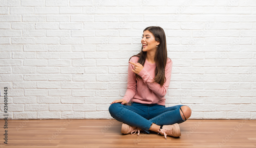 Teenager girl sitting on the floor in a room pointing to the side to present a product Teenager girl sitting on the floor in a room pointing to the side to present a product