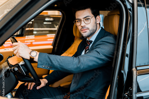 stylish businessman in eyeglasses looking at camera while sitting in luxury car