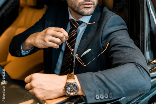 partial view of businessman with luxury watch holding eyeglasses while sitting in car photo