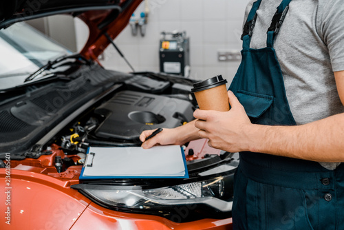partial view of male auto mechanic with notepad and coffee to go near car at auto repair shop