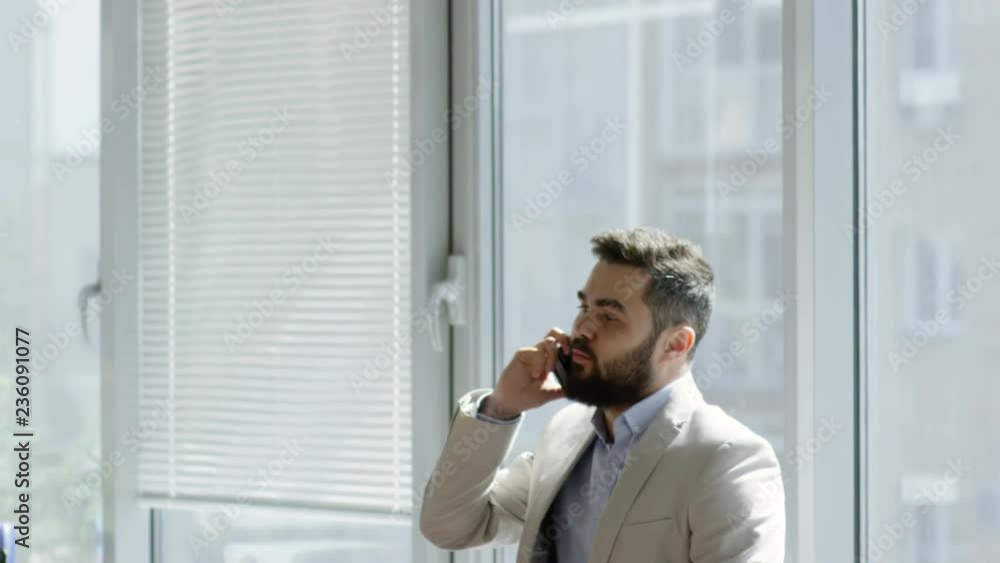 Tilt down of bearded Caucasian businessman standing by the window in office and talking on mobile phone Tilt down of bearded Caucasian businessman standing by the window in office and talking on mobile phone