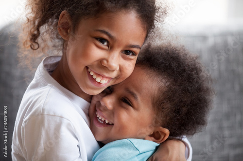 Happy African American siblings embracing, sitting together on couch at home in living room, little preschooler girl hug toddler adorable boy, good relations between sister and brother, close up