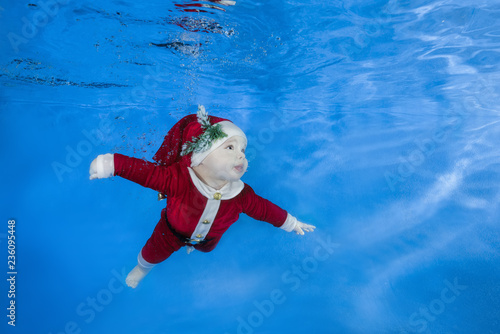 Little baby boy dressed as Santa swim underwater. Baby swimming underwater in the pool on a blue water background. Healthy family lifestyle and children water sports activity.