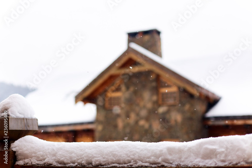 Snow-covered surface for product placement. Blurred house made of wood and stone on background. Snowfall on winter day. Christmas background with wooden cottage or chalet at ski resort. Copyspace.