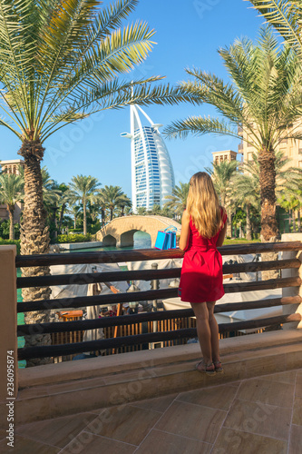 Fotografie, Obraz Young tourist woman dressed in red dress looking Burj al Arab from Madinat Jumeirah souk with shopping bags next to her