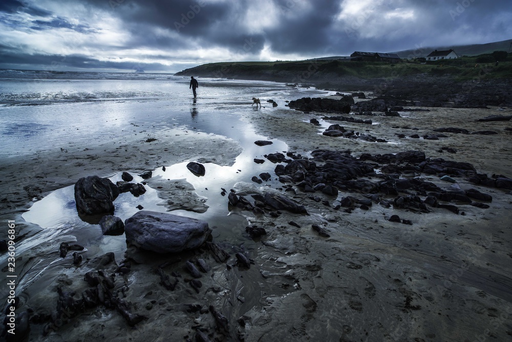 Obraz premium Atmospheric scene of a man and dog on a beach in Ireland in front of Skelly Island with an approaching storm with dramatic dark threatening clouds