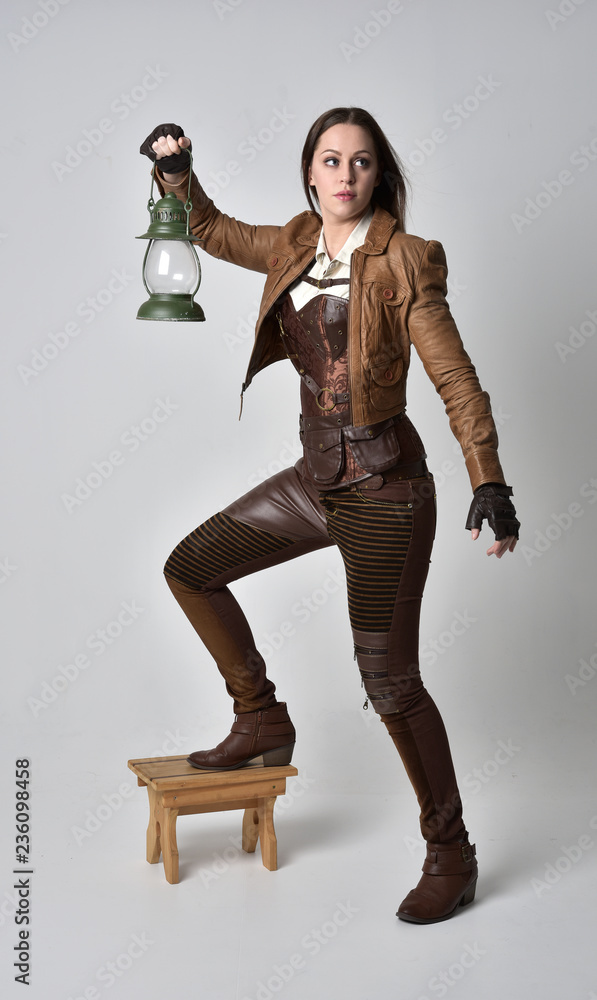 full length portrait of brunette girl wearing brown leather steampunk outfit. standing pose, holding a gas lantern, on grey studio background. full length portrait of brunette girl wearing brown leather steampunk outfit. standing pose, holding a gas lantern, on grey studio background.