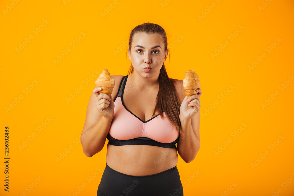Image of amusing chubby woman in tracksuit holding two croissants in both hands, isolated over yellow background Image of amusing chubby woman in tracksuit holding two croissants in both hands, isolated over yellow background