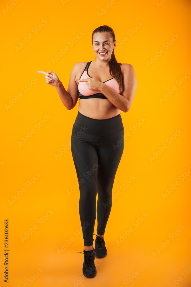 Full length portrait of big woman in sportive bra smiling and pointing finger aside, isolated over yellow background Full length portrait of big woman in sportive bra smiling and pointing finger aside, isolated over yellow background