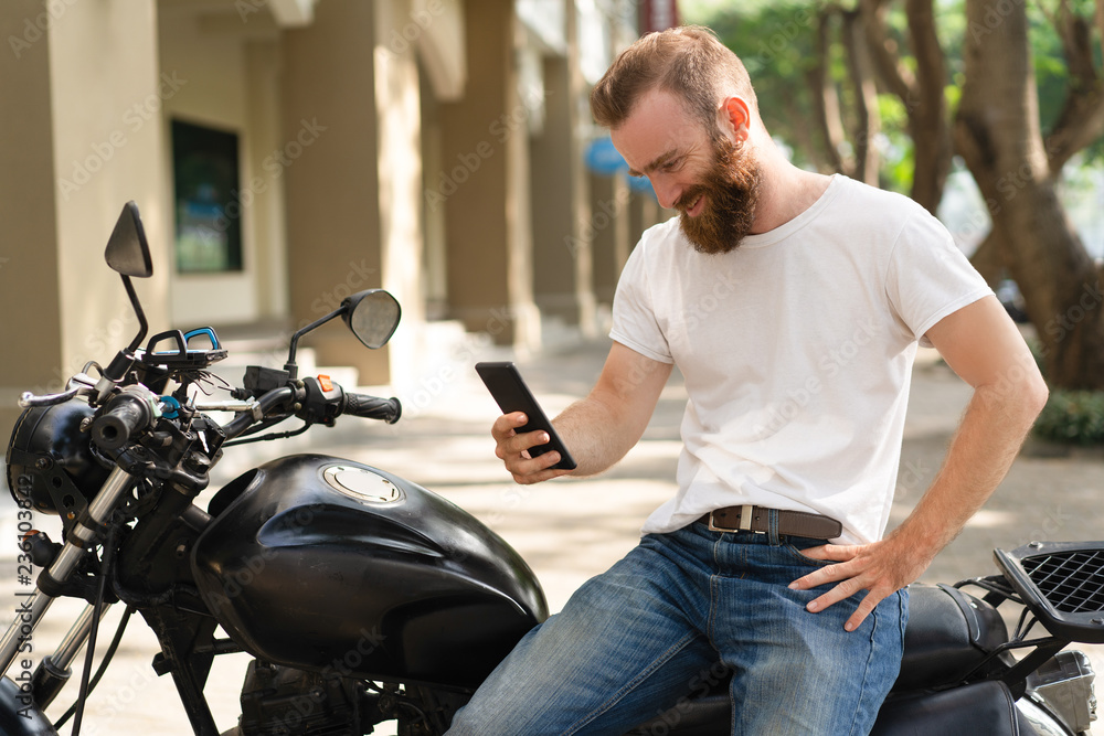 Cheerful motorbike rider having video call. Young man with bushy beard sitting on motorcycle and smiling at phone screen. Communication concept