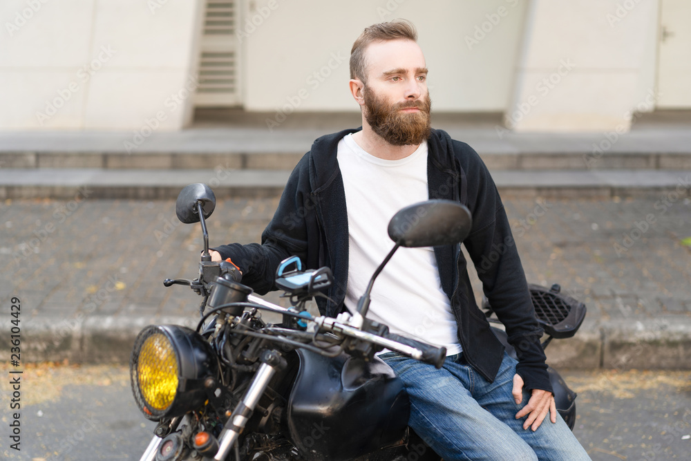 Pensive biker waiting girlfriend at parking space. Serious young man with beard sitting on motorbike and looking into distance. Riding bike or waiting concept Pensive biker waiting girlfriend at parking space. Serious young man with beard sitting on motorbike and looking into distance. Riding bike or waiting concept