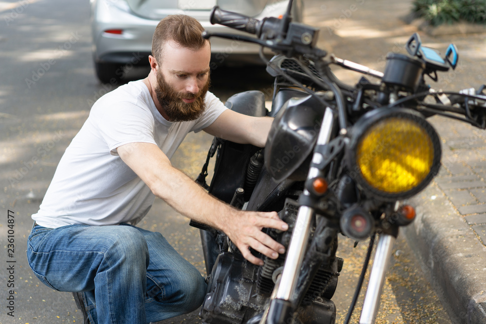 Serious biker diagnosing broken motorbike. Bearded young man sitting on haunches and examining motorcycle. Breakdown concept