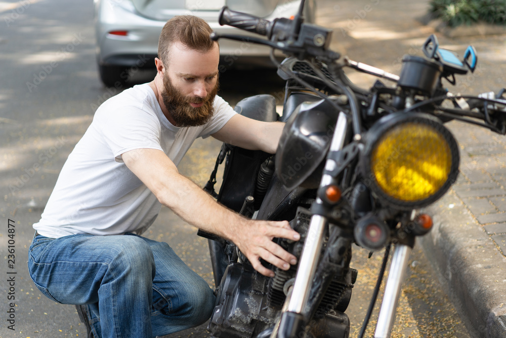 Obraz premium Serious biker diagnosing broken motorbike. Bearded young man sitting on haunches and examining motorcycle. Breakdown concept