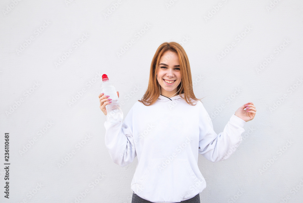 Portrait of a happy, attractive girl in sportswear and a bottle of water in her hands, listens to music in the headphones, looks at the camera and smiles. Joyful sports girl on a white background Portrait of a happy, attractive girl in sportswear and a bottle of water in her hands, listens to music in the headphones, looks at the camera and smiles. Joyful sports girl on a white background