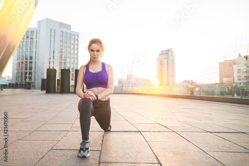 Portrait of a young sports girl squatting against the background of the sunset. Girl in sports wear sports on the street. Sports training in the background of the sunrise. Sports concept.