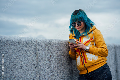 Portrait of young woman with dyed blue hair leaning against wall looking at cell phone photo