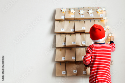 Little baby Santa's helper is watching the advent calendar for Christmas. Mood Board with gifts and star garland .