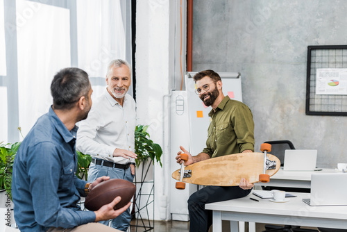 handsome businessmen talking and holding skateboard and american football ball in office