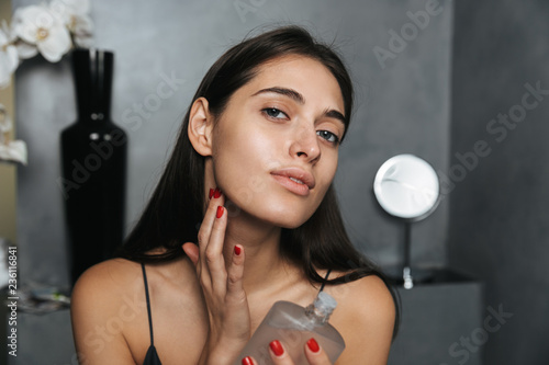 Photo of brunette woman with long dark hair standing in bathroom, and holding bottle with perfume