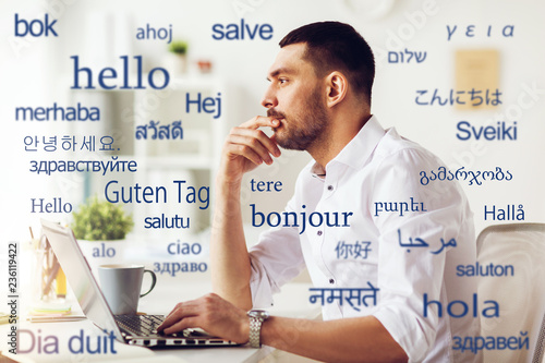 translation, business, and technology concept - male translator or businessman with laptop computer thinking at office over greeting words in different foreign languages photo