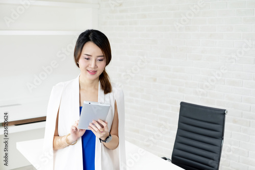 Portrait of beautiful Asian business woman in white suit and blue dress standing in front of the table and using a tablet