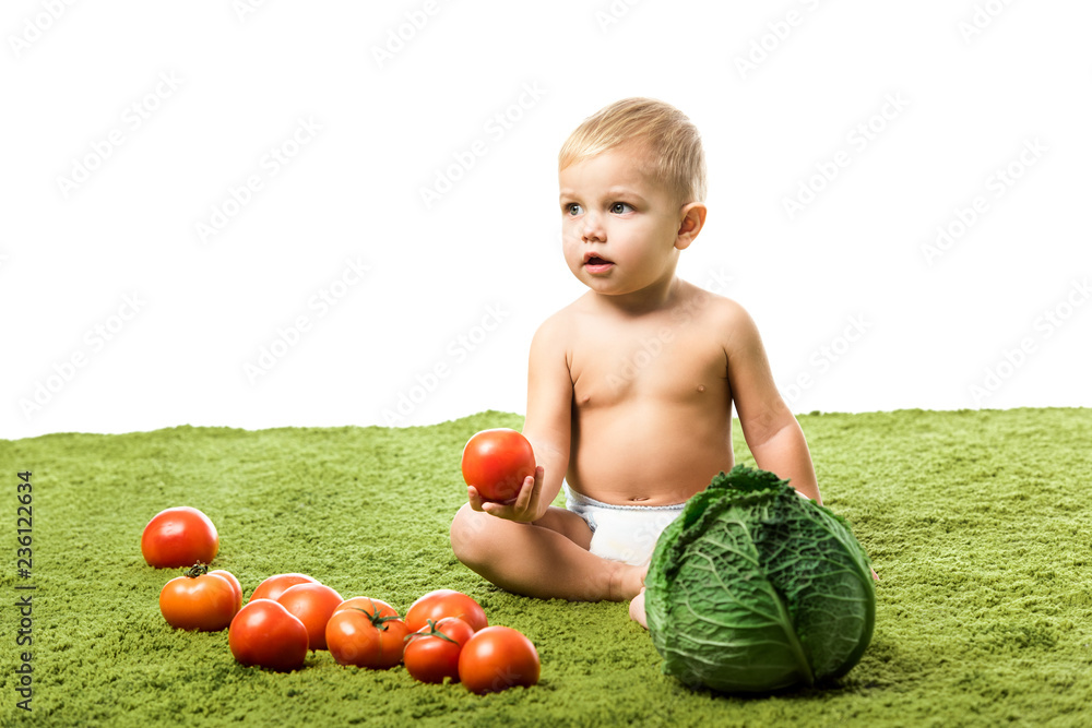 Fototapeta premium adorable toddler boy holding tomato and sitting on green carpet with vegetables isolated on white