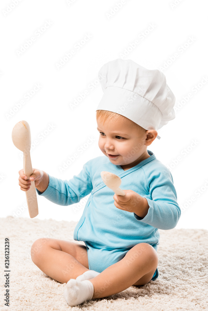 toddler boy in chefs hat sitting on carpet, holding two big wooden spoons and smiling isolated on white toddler boy in chefs hat sitting on carpet, holding two big wooden spoons and smiling isolated on white