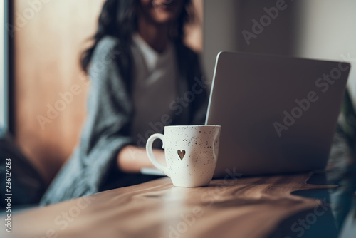 Laconic image of white cup with tiny heart on it standing on the table near modern laptop