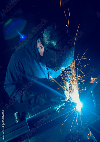 A vibrant action shot of a skilled working metal welder in action, welding metal. Photographed with a slow shutter speed and spark trails. Orange and teal.