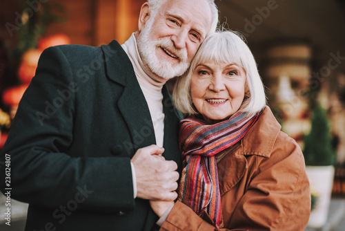 Portrait of happy bearded gentleman hugging his lovely wife and holding her hand. They looking at camera and smiling