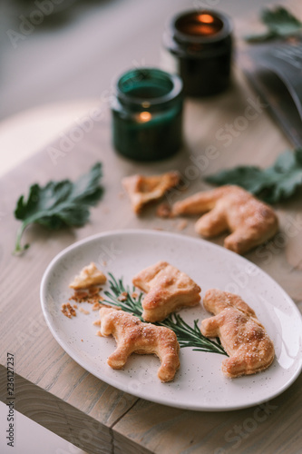 Homemade shaped cookies on a wooden table next to candles, green leaves and a cookbook. Crumbs are scattered at ease on the table. The concept of Christmas atmosphere and comfort.