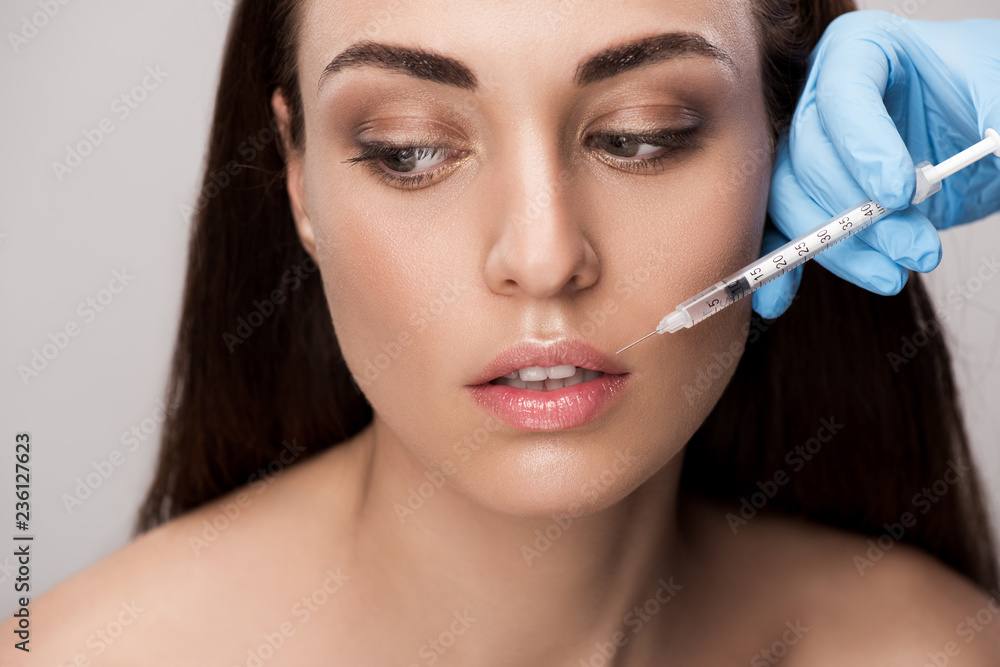 doctor making beauty injection into lips for attractive girl isolated on grey doctor making beauty injection into lips for attractive girl isolated on grey
