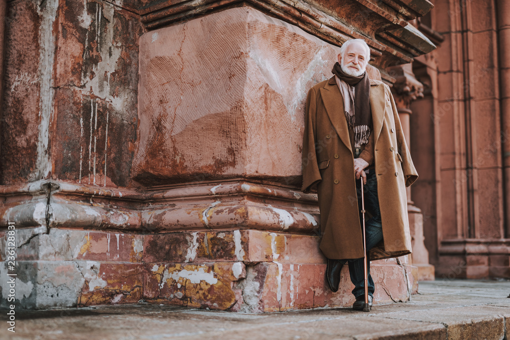 Full length portrait of senior bearded gentleman in coat standing on the street. He is looking at camera and smiling