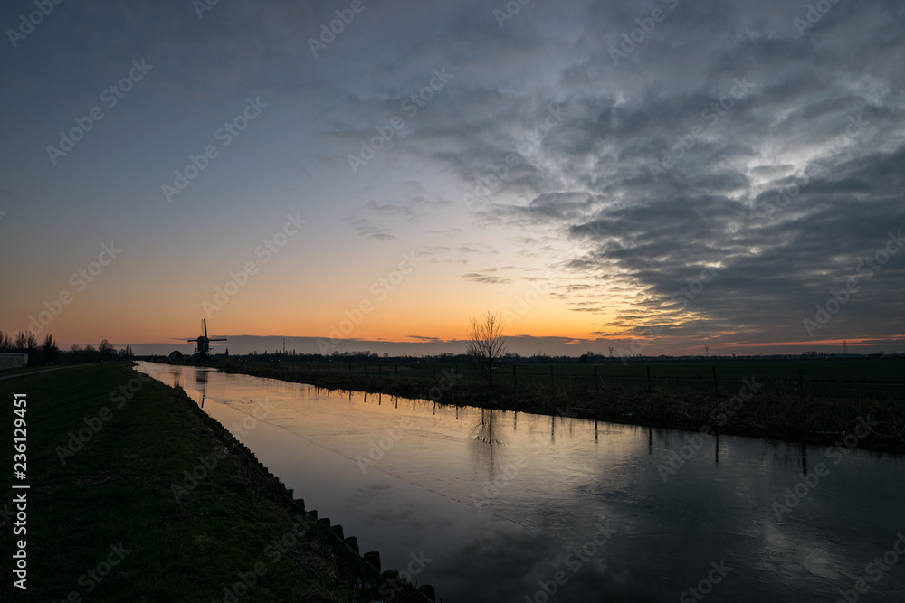 A dutch canal is covered with ice. A distant windmill is silhouetted against the evening sky.